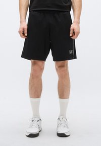 Čierne športové šortky z priedušnej tkaniny, s elastickým pásom a logom. Nosené s bielymi ponožkami a športovou obuvou.