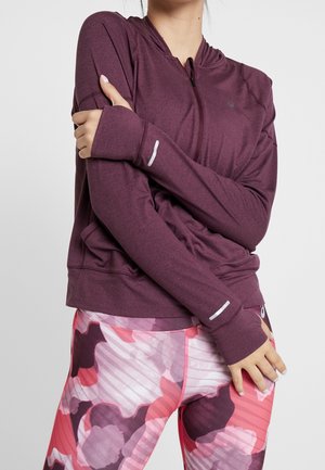 Frau trägt eine bordeauxrote Sportjacke mit Reißverschluss und Daumenlöchern sowie rosa Leggings mit Blumenmuster, die Arme vor der Brust verschränkt.