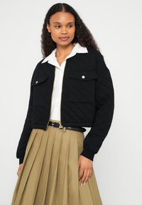 Μαύρο κοντό καπιτονέ μπουφάν με χρυσά κουμπιά τύπου σναπ και δύο μπροστινές τσέπες, συνδυασμένο με λευκό πουκάμισο και μπεζ φούστα με πτυχώσεις.