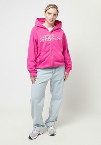 Sudadera rosa con cremallera y un logo blanco, de mangas largas y capucha con cordones, combinada con jeans azul claro y zapatillas deportivas grises.
