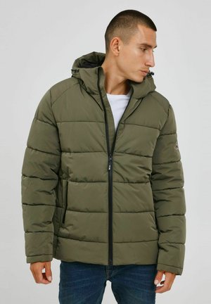 Doudoune verte olive avec capuche, texture matelassée, fermeture éclair à l'avant et deux poches latérales. Portée sur un t-shirt blanc et un jean bleu.