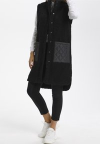 Gilet nero, fluffy e senza maniche con chiusura a bottoni, tessuto texturizzato e due tasche grigie trapuntate. Indossato sopra una maglietta bianca a maniche lunghe.
