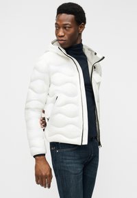 Άνδρας που φοράει λευκό μπουφάν με επένδυση και κουκούλα, μπλε ζιβάγκο και σκούρο μπλε τζιν, κοιτώντας προς τα αριστερά του.