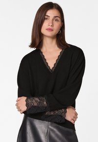 Schwarzer V-Ausschnitt-Strickpullover mit Spitzenbesatz an den Ärmelbündchen und dem Ausschnitt, lockere Passform und strukturierter Stoff.