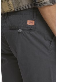 Pantaloni gri închis cu o textură netedă, având un buzunar posterior, închidere cu nasture și o etichetă din piele cu numele brandului.