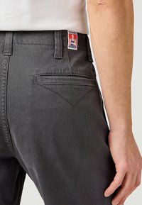 I pantaloni in denim grigio presentano un design a gamba dritta, con una tasca posteriore che mostra un dettaglio a punto V. L'etichetta sulla cintura reca la scritta "Wrangler".