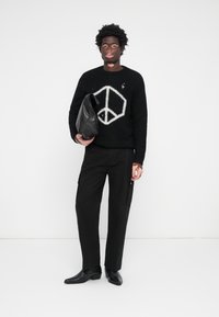 Pulover negru din tricot cu un semn de pace alb, pantaloni cargo negri și pantofi negri. Ține o geantă din piele neagră. Fundal simplu.