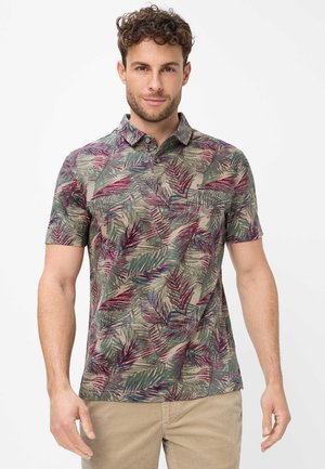 Man draagt een poloshirt met korte mouwen met een meerkleurig tropisch bladerenpatroon en een beige broek, staand tegen een witte achtergrond.