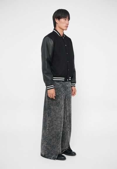 Giacca varsity nera con maniche in pelle, polsini a righe, abbinata a pantaloni di jeans grigi lunghi e strappati con uno spacco frontale. Scarpe nere.