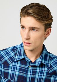 Camicia a quadri blu e verde acqua con chiusura a bottoni, colletto strutturato e due tasche sul petto; il tessuto appare texturizzato e resistente.