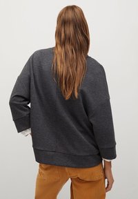 Sweatshirt oversize gris foncé avec une coupe ample, épaules tombantes et larges manches, superposé sur une chemise blanche et associé à un pantalon beige.