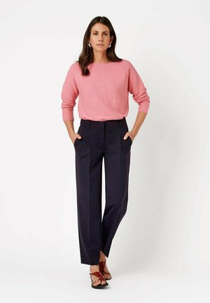 Woman with dark hair wearing a pink long-sleeve sweater, dark tailored pants, and brown open-toe heels, standing with hands in pockets.