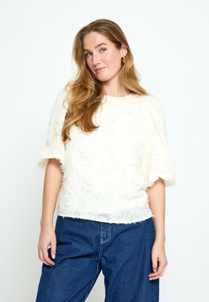 Kobieta o długich falowanych włosach, nosząca złote kolczyki-koła, białą teksturowaną bluzkę z krótkim rękawem oraz luźne niebieskie jeansy, stoi na białym tle.