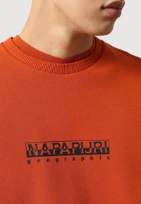 Sudadera naranja con cuello acanalado, con un logo negro impreso que dice "NAPAPIJRI geographic" centrado en la parte frontal. Textura suave.