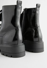 Bottines en cuir verni noir avec semelles épaisses, fermetures à glissière latérales et tirants en boucle. Texture lisse avec une finition brillante.