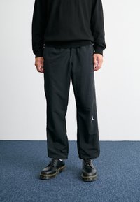 Black athletic pants made of lightweight fabric; feature a loose fit and a white Jordan logo on the left leg. Paired with black boots.