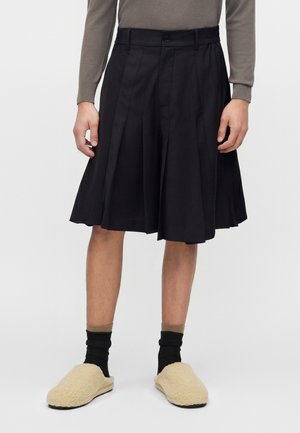 Osoba oblečená v čiernych plisovaných kraťasoch po kolená, béžových nazúvacích papučiach, čiernych ponožkách a dlhom rukáve taupe topu, stojaca na jednoduchom pozadí.