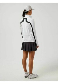 Top bianco a maniche lunghe con linee nere, abbinato a una gonna nera a pieghe. Un cappello argento e delle scarpe bianche completano l'outfit.