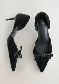 Para czarnych aksamitnych szpilek z wąskim noskiem z paskami na kostkę i małymi kokardkami na jasnym tle.