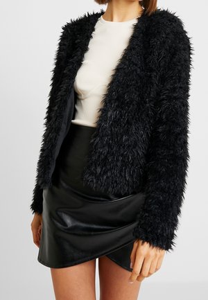 Mujer que lleva una chaqueta negra con textura peluda sobre un top blanco y una minifalda de cuero negra con detalle de envoltura.