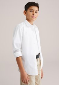 Cămașă albă cu nasturi, mâneci lungi rulate la manșete. Purtată peste pantaloni de culoare crem, având o curea împletită neagră.