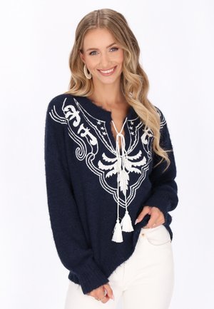 Granatowy sweter dzianinowy z białymi haftowanymi wzorami, otwarty dekolt z wiązaniem i frędzlami, długie rękawy i luźny krój.