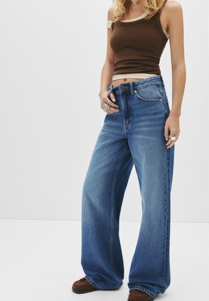 Jeans en denim bleu à jambes larges avec un lavage délavé, comportant cinq poches et une fermeture par bouton. Associés à un débardeur en maille marron.