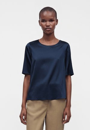 Femme portant un haut en satin bleu foncé à manches courtes et un pantalon beige, debout devant un fond clair uni.