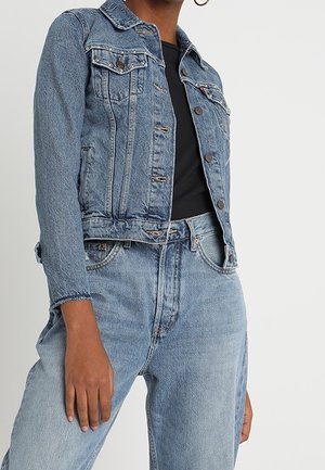 Person wearing light blue denim jacket and matching high-waisted jeans with a black top, against a plain white background.