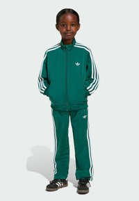 Malo dijete stoji, odjeveno u zeleni Adidas trenirku s bijelim prugama i crnim Adidas tenisicama, ruke u džepovima jakne.