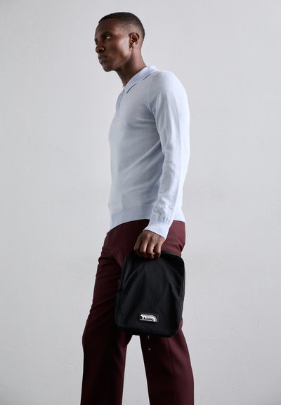 Polo shirt bleu clair à manches longues avec des poignets côtelés, associé à un pantalon bordeaux. Tient un sac à dos noir avec un patch logo.