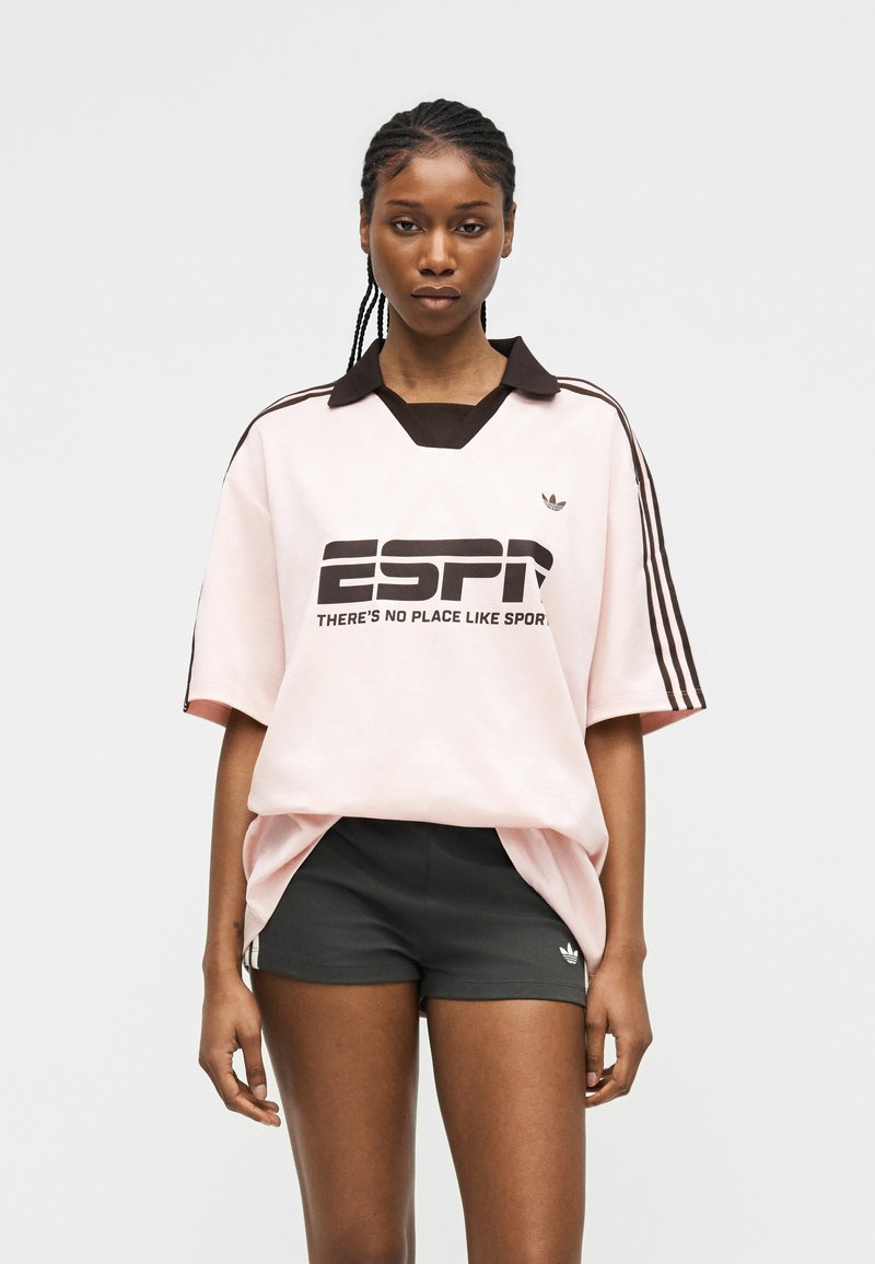 Femme aux cheveux tressés portant un t-shirt de sport Adidas ESPN rose clair et un short Adidas noir, debout devant un fond gris uni.