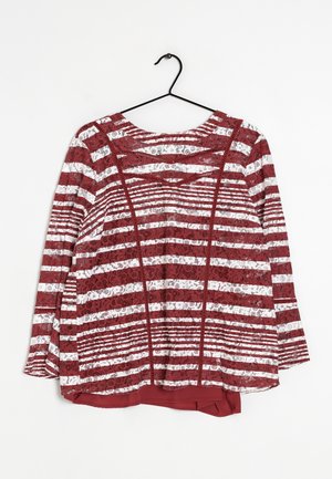 Blouse en dentelle à manches longues rayée rouge et blanche avec motif floral, suspendue sur un cintre noir contre un fond blanc.