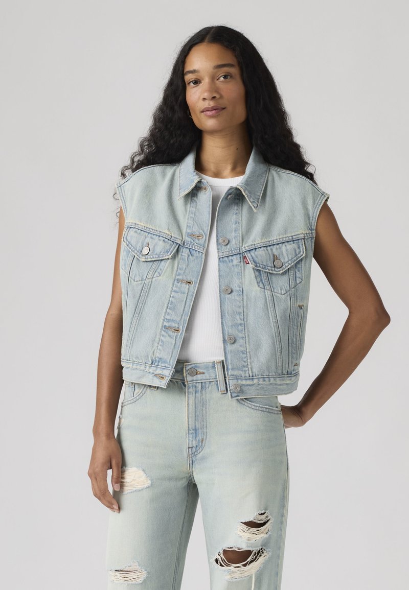 Femme aux longs cheveux bouclés portant un gilet en denim bleu clair sans manches et un jean bleu clair déchiré, debout avec une main sur la hanche.