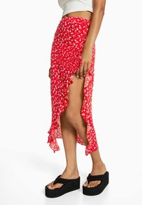 Punainen kukkainen hame, jossa on röyhelöreuna ja korkea halkio, valmistettu kevyestä kankaasta. Yhdistetty mustien alustohanskojen kanssa, joissa on leveät remmit.