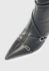 Botín negro de cuero hasta el tobillo con punta afilada, que presenta correas cruzadas y un detalle de hebilla plateada. Textura suave y diseño estructurado.