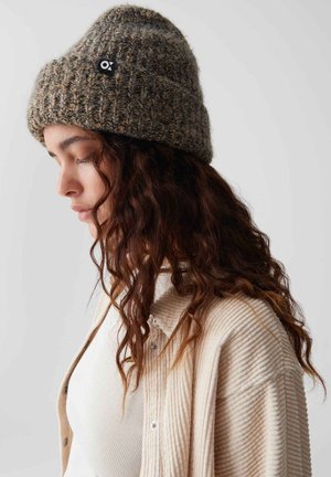 Beanie marrone in maglia con un motivo texturizzato e una piccola toppa nera con logo. Il modello indossa un top a coste crema e una camicia in velluto a coste.