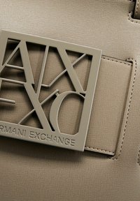 Borsa a mano in pelle sintetica beige con una placca logo geometrica, lettering stampato in rilievo e dettagli di cucitura in vista.