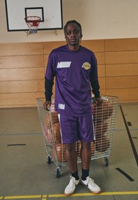 Uniforme de baloncesto morado con textura de malla, que presenta el logo de la NBA y el texto "living". Acompañado de una canasta de alambre llena de balones de baloncesto.