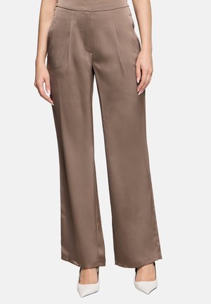 Woman wearing high-waist, wide-leg brown satin trousers paired with white pointed-toe heels, standing against a plain background.