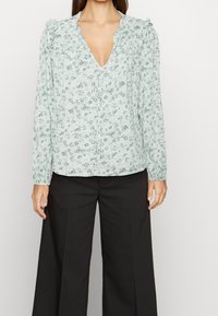 Blouse florale vert clair avec un décolleté en V, des manches longues, des accents d'épaule froncés et une fermeture à boutons ; portée avec un pantalon large noir.