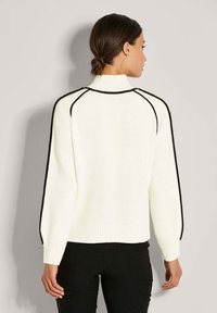 Frau mit dunklem Haar in einem tiefen Haarknoten, die einen weißen Strickpullover mit schwarzem Rand an den Raglanärmeln und schwarze Hosen trägt, mit dem Rücken zur Kamera.