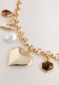 Gold-tone metal necklace featuring two heart-shaped charms, a pearl, and a hexagonal charm, with a linked chain design and shiny finish.