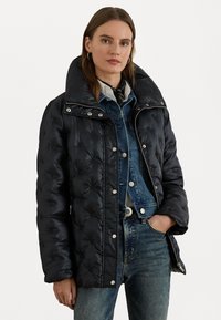 Black puffer jacket with a wide collar, quilted texture, and silver snap fasteners. Worn over a denim jacket and paired with blue jeans.