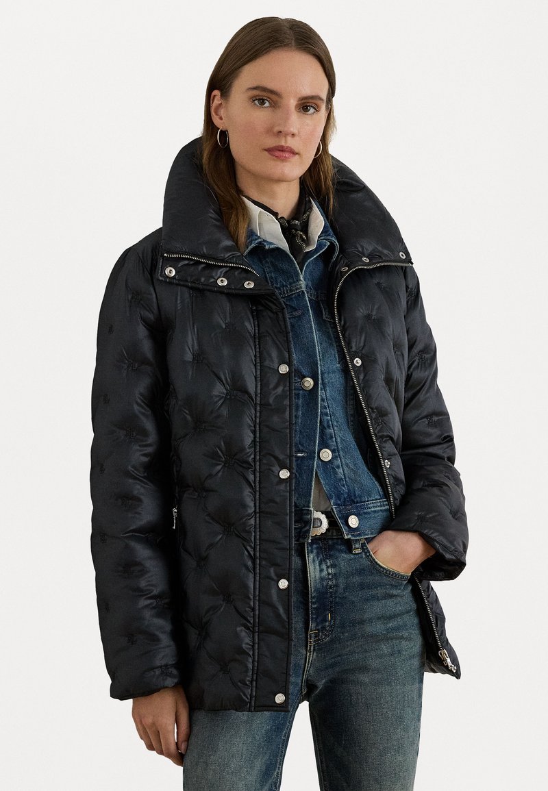 Black puffer jacket with a wide collar, quilted texture, and silver snap fasteners. Worn over a denim jacket and paired with blue jeans.