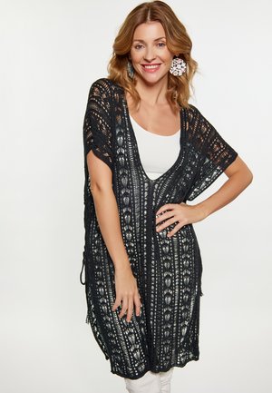 Donna sorridente che indossa grandi orecchini floreali, una maglietta bianca e un lungo cardigan nero a maglia aperta con maniche corte.