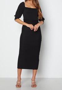 Zwarte geribbelde midi-jurk met een vierkante halslijn en pofmouwen. Gecombineerd met doorzichtige, hoge sandalen met een kralenband.
