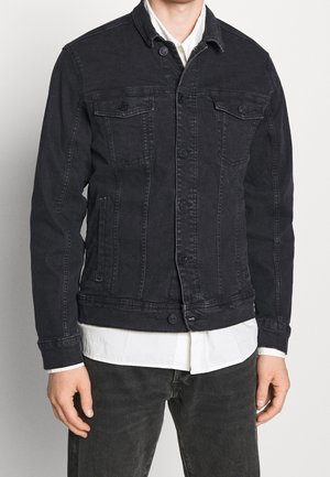 Man draagt een zwarte spijkerjasje over een wit shirt met zwarte jeans, staand tegen een effen lichte achtergrond.