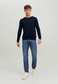 Tengerészkék kötött pulóver hosszú ujjakkal, kerek nyakkal, világoskék farmerszerű nadrággal és fehér sportcipővel, világos háttér előtt állva.