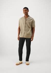 Short-sleeve, khaki button-up shirt in a textured fabric, paired with black trousers and white sneakers with tan accents.